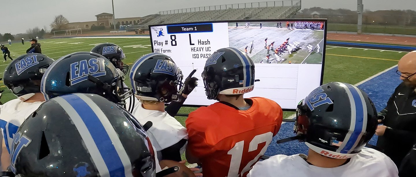 a group of athletes grouped in a field, watching the video of a previous match on a Bretford's heavy duty tv cart