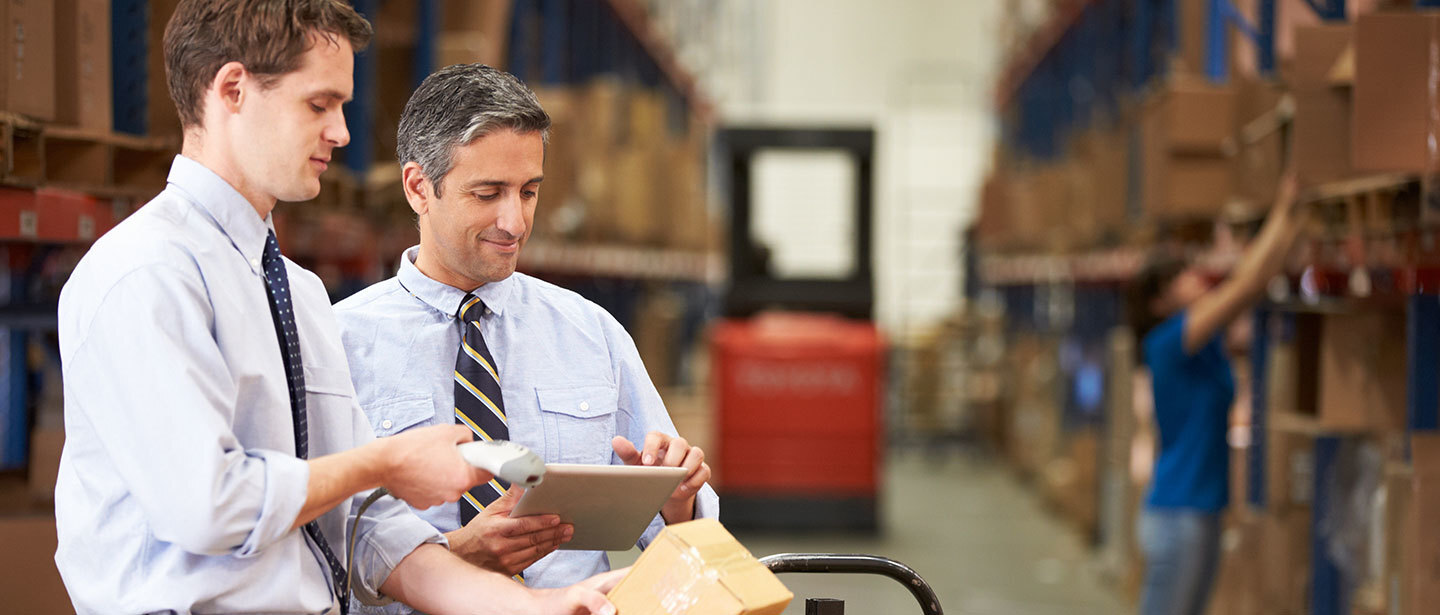 two warehouse managers reviewing the details of a product using a tablet, while some employees are working in the background